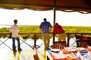 A wooden deck with a table set for dining, overlooking a body of water. - Camp Linyanti (Chobe National Park)