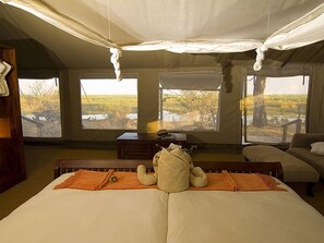 A bedroom with a large bed, a wooden bench, and a window overlooking a natural landscape. - Camp Linyanti (Chobe National Park)
