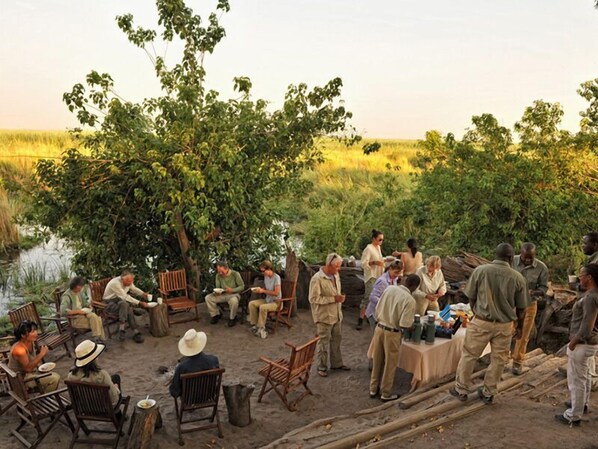 A group of people seated on chairs and benches in an outdoor setting with trees and a body of water in the background. - Camp Linyanti (Chobe National Park)