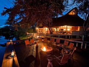 An outdoor dining area with a fire pit, wooden deck, and seating. - Camp Linyanti (Chobe National Park)