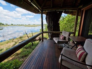 A wooden deck with two chairs and a table, overlooking a calm lake. - Camp Linyanti (Chobe National Park)