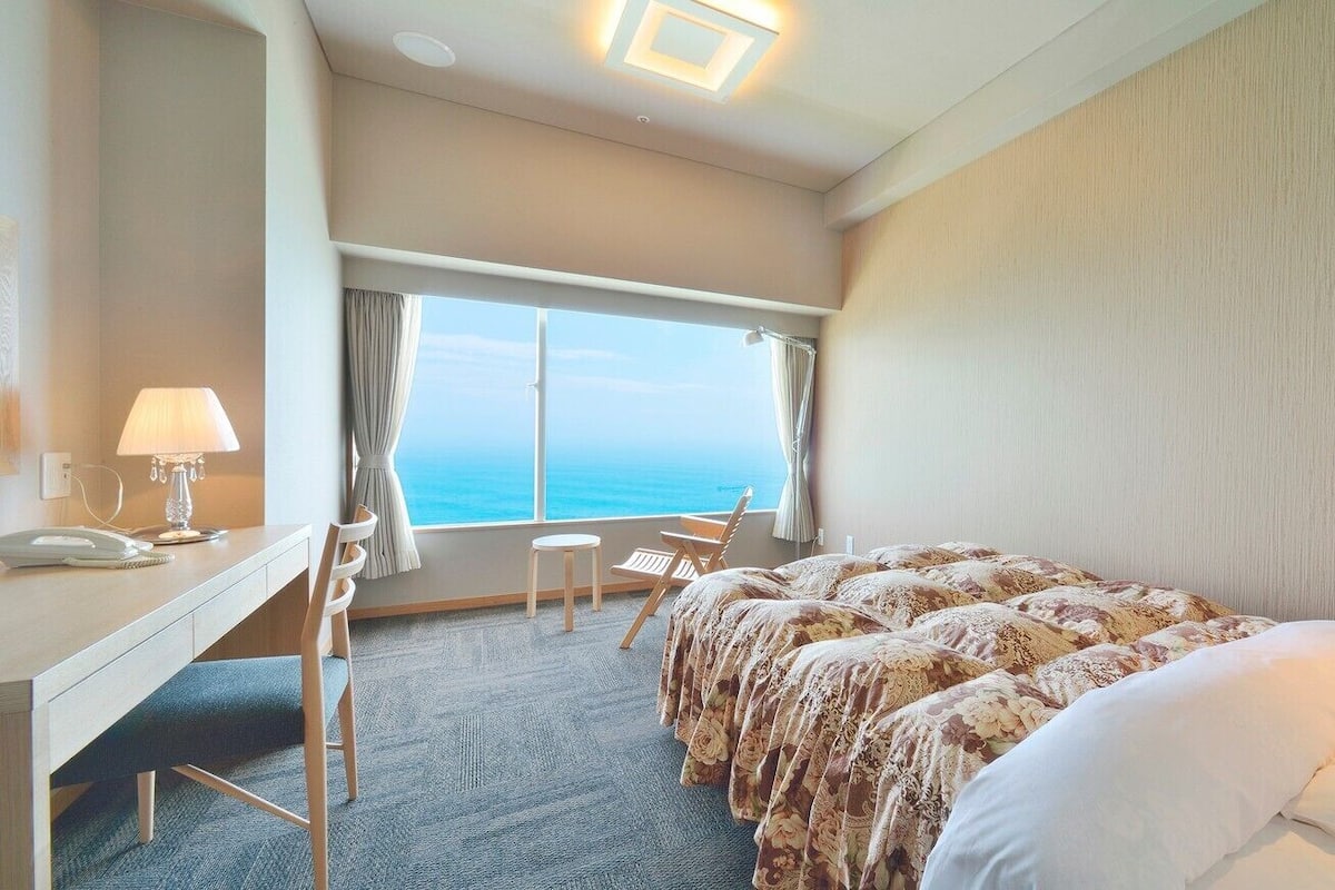 Ein Hotelzimmer mit Bett, Schreibtisch, Stuhl, Lampe und Blick auf den Ozean.