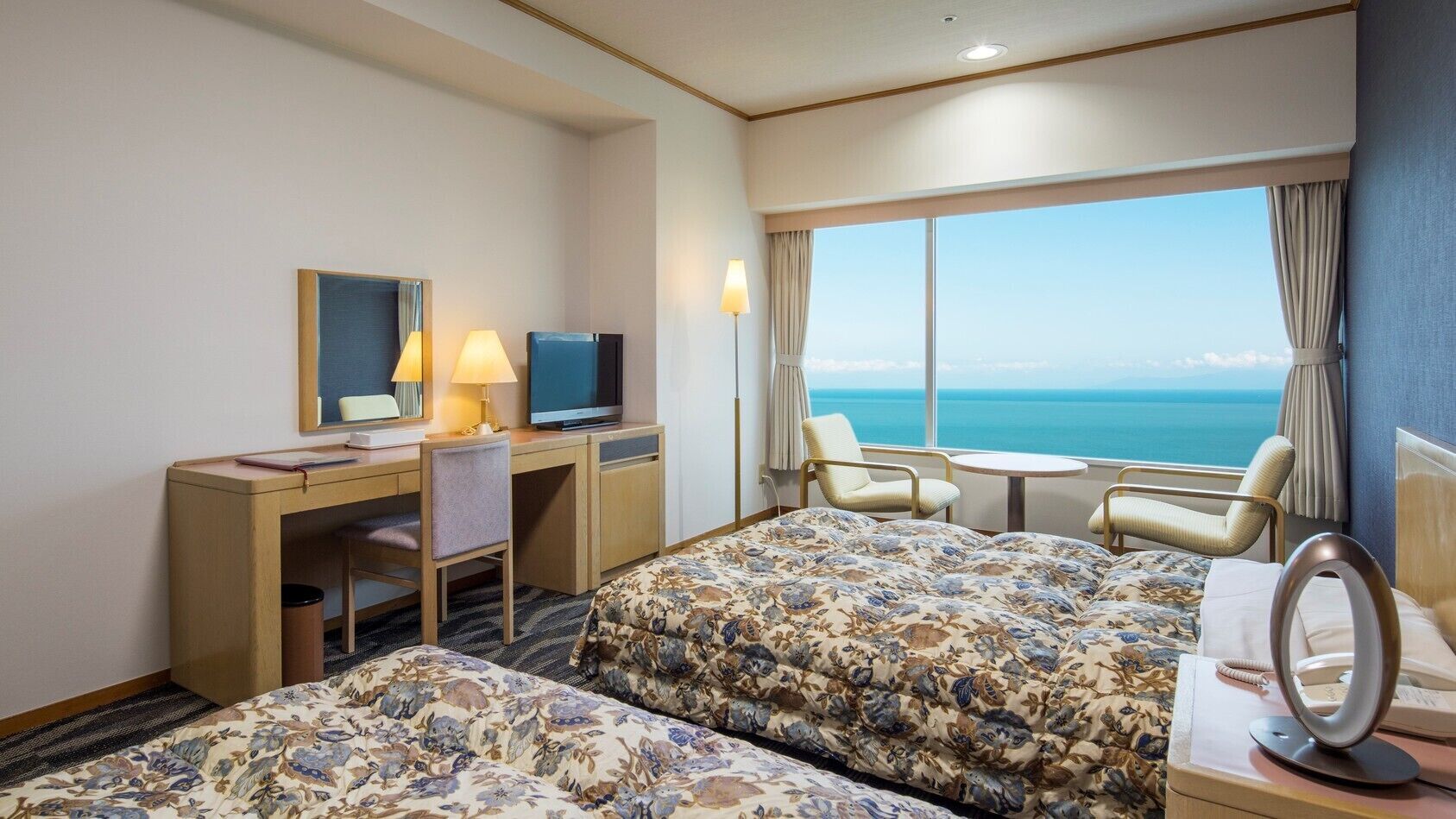 Ein Hotelzimmer mit Bett, Schreibtisch mit Computer, Stuhl, kleinem Tisch und Meerblick.
