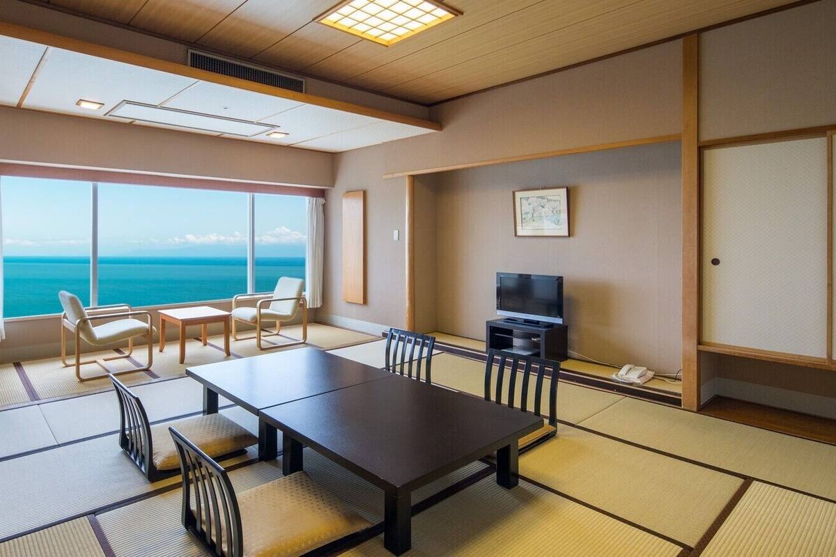 Une pièce dotée d’un sol recouvert de tatamis, d’une table à manger, de chaises, d’un téléviseur et d’une grande fenêtre donnant sur la mer.
