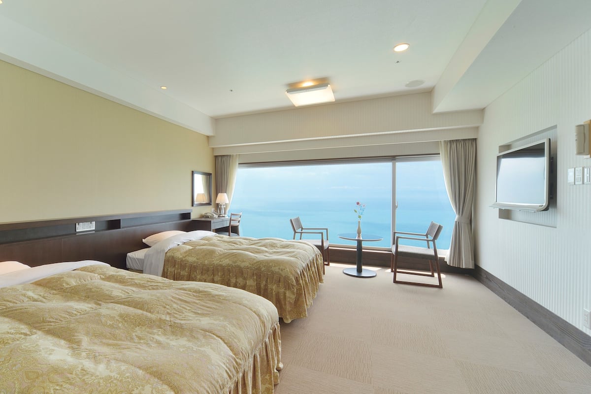 Ein Hotelzimmer mit zwei Betten, einem Schreibtisch, einem Sessel, einem Fernseher und Meerblick.