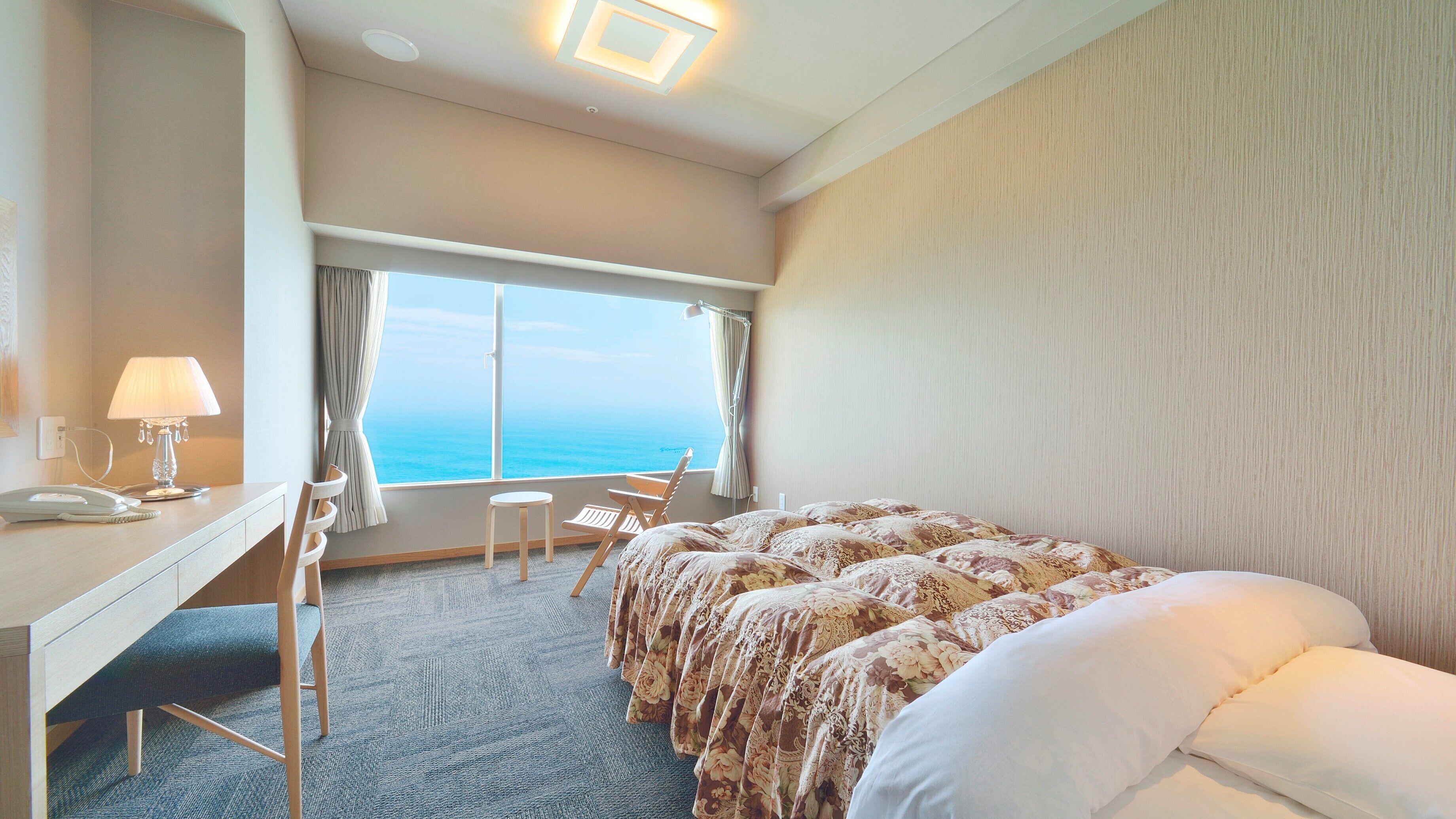 Ein Hotelzimmer mit Bett, Schreibtisch, Stuhl, Lampe und einem großen Fenster mit Meerblick.