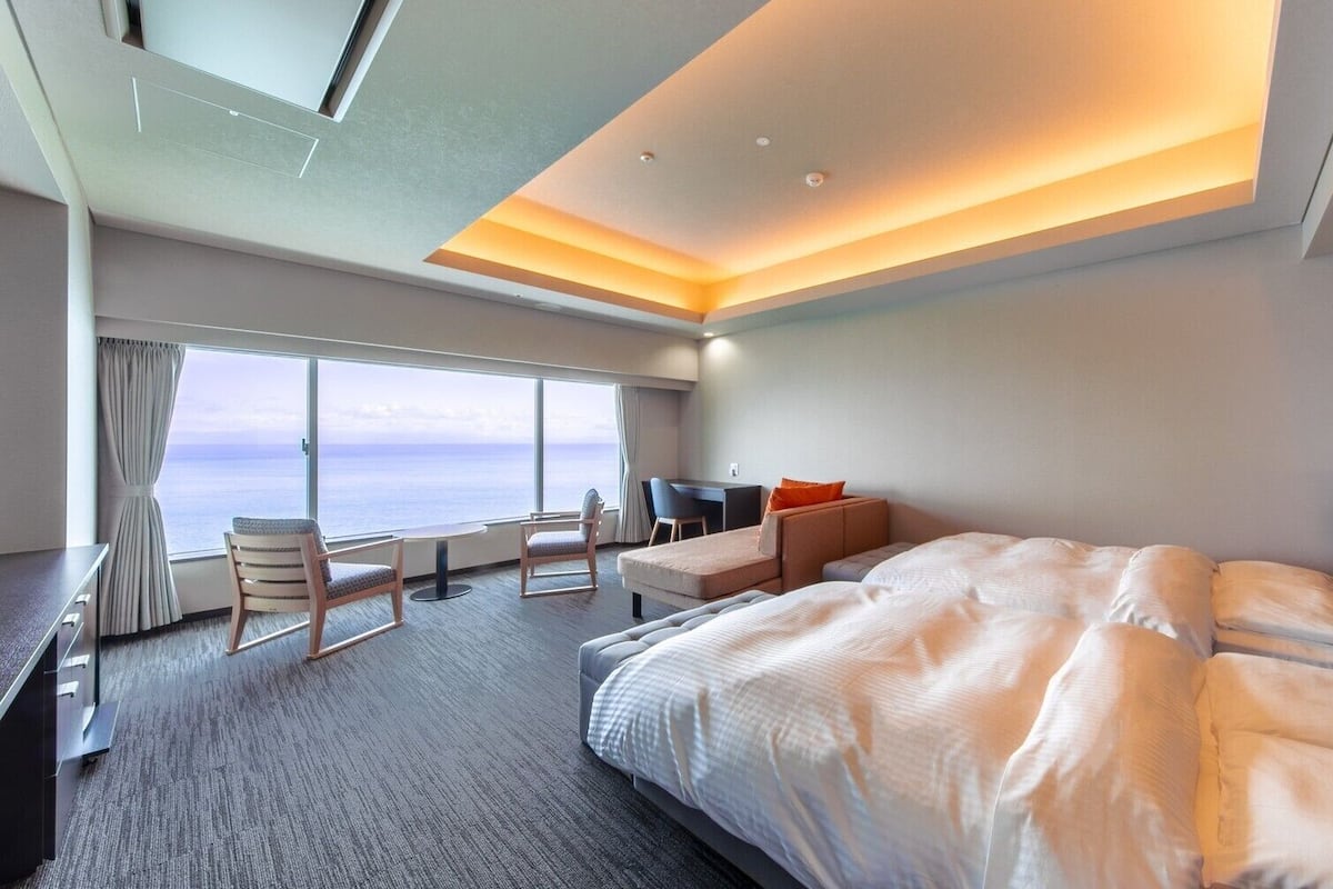 Ein modernes Hotelzimmer mit einem großen Bett, einem Schreibtisch, einem Sessel und Blick aufs Meer.
