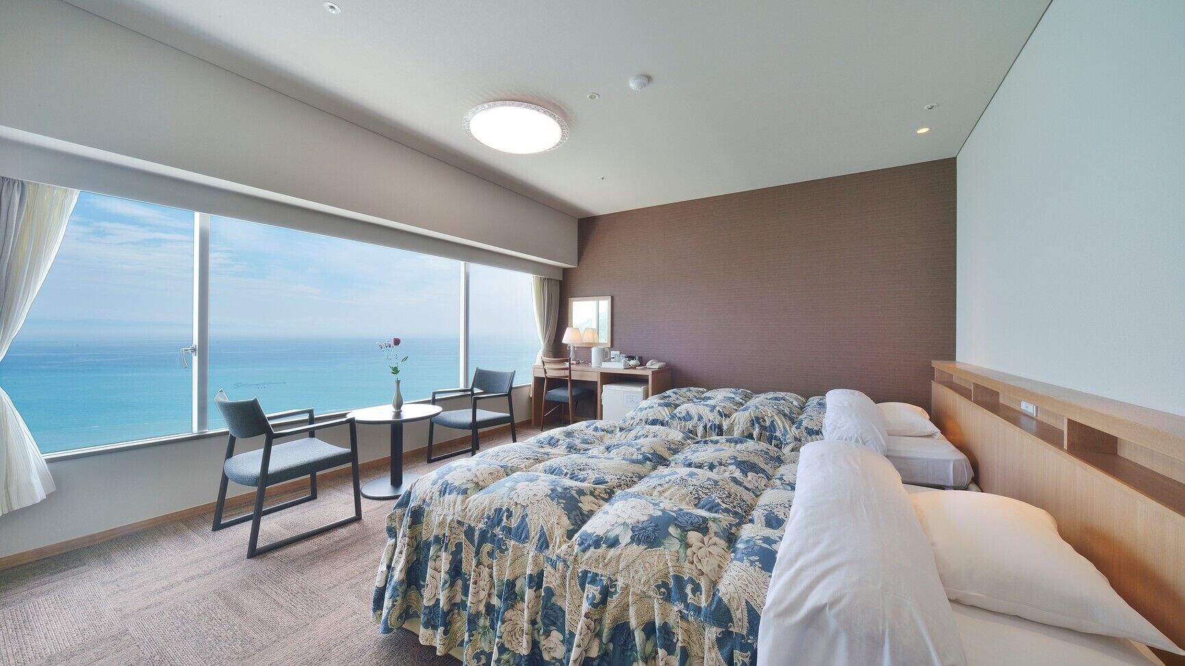 Ein Hotelzimmer mit einem großen Bett, einem Schreibtisch und einem Stuhl. Durch große Fenster ist der Blick auf das Meer.