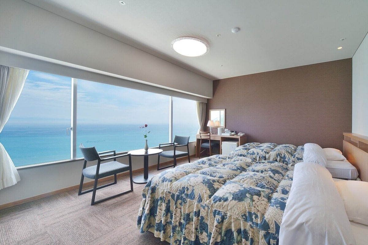 Ein Hotelzimmer mit einem großen Bett, einem Schreibtisch und einem Stuhl. Durch große Fenster ist der Blick auf das Meer.