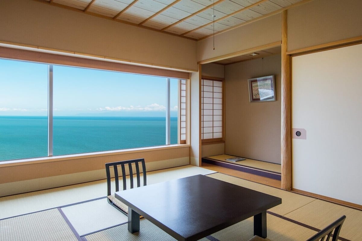 Ein Zimmer mit Tatami-Boden, einem niedrigen Tisch, einem Stuhl, einer Schiebetür und Meerblick.