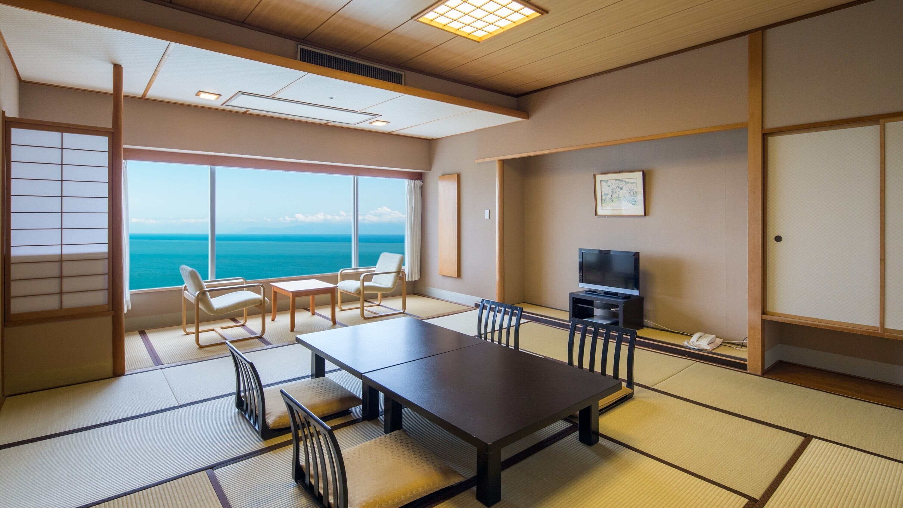 Ein Zimmer mit Tatami-Boden, einem Fernseher, einem Tisch, Stühlen und Blick auf den Ozean.