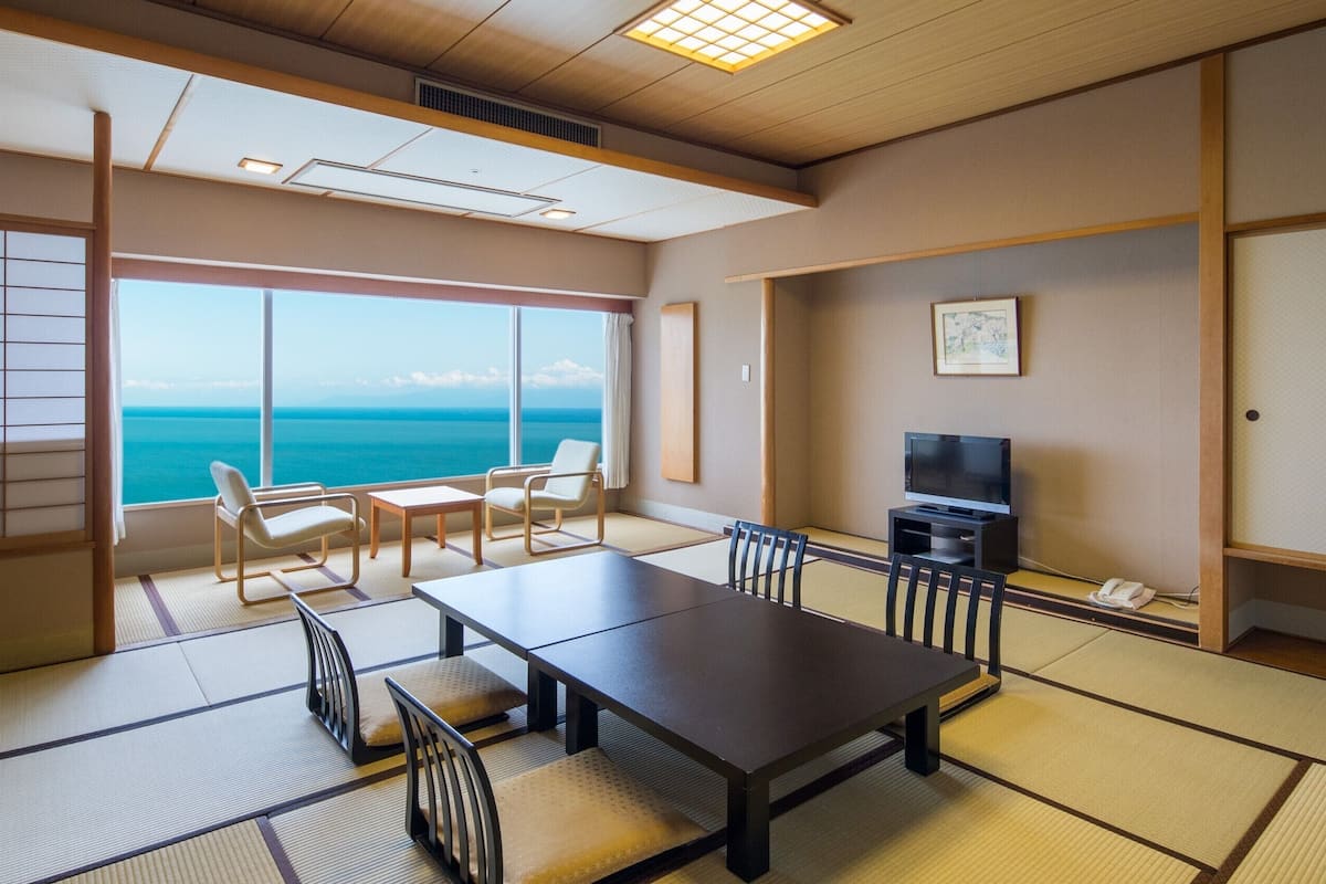 Ein Zimmer mit Tatami-Boden, einem Fernseher, einem Tisch, Stühlen und Blick auf den Ozean.