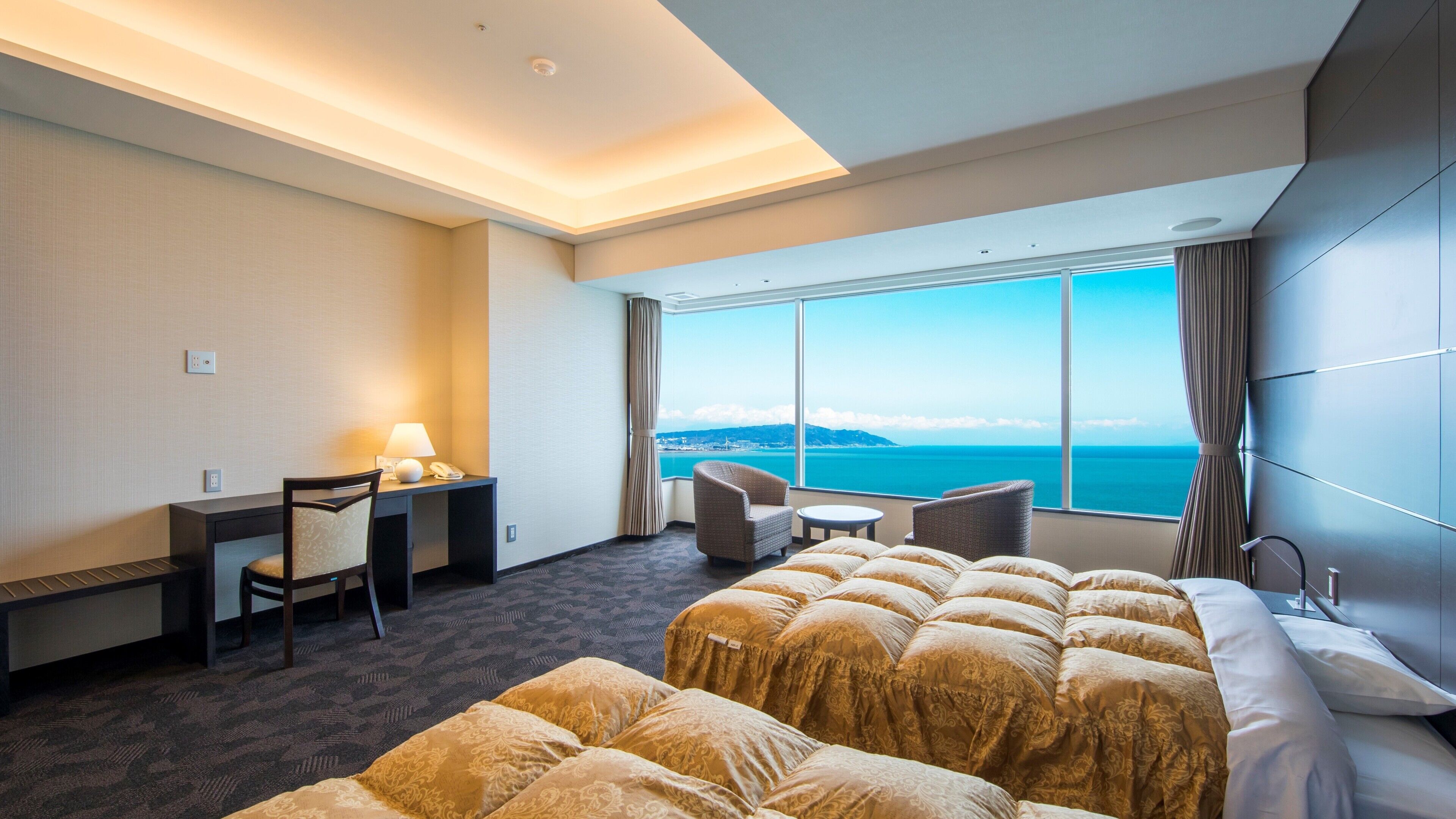 Ein Hotelzimmer mit Bett, Schreibtisch mit Lampe, Stuhl, kleinem Tisch und Meerblick.