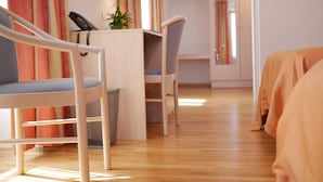 Hotellihuone, jossa on puulattia, kirjoituspöytä lampun kanssa, tuoli ja sänky, jonka päällä on oranssi peitto.