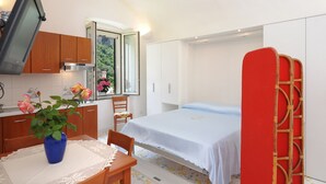 Una habitación compacta con cama, una mesita con un florero, una ventana con vistas al verdor y una kitchenette con armarios de madera y televisión.