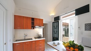 Una cocina con armarios de madera, encimera blanca y salpicadero de azulejos. Se observa un fregadero, una estufa y una ventana con vistas al exterior.