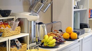 Una encimera de cocina con una cafetera, un hervidor y diversos alimentos como plátanos, uvas y naranjas.