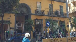 Un edificio amarillo de varios pisos con balcones, una calle delante y un coche azul estacionado cerca.