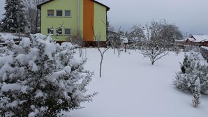 A two-story building with a green and orange facade, surrounded by snow-covered trees and shrubs. - Gästezimmer Vogelsberg (Freiensteinau)