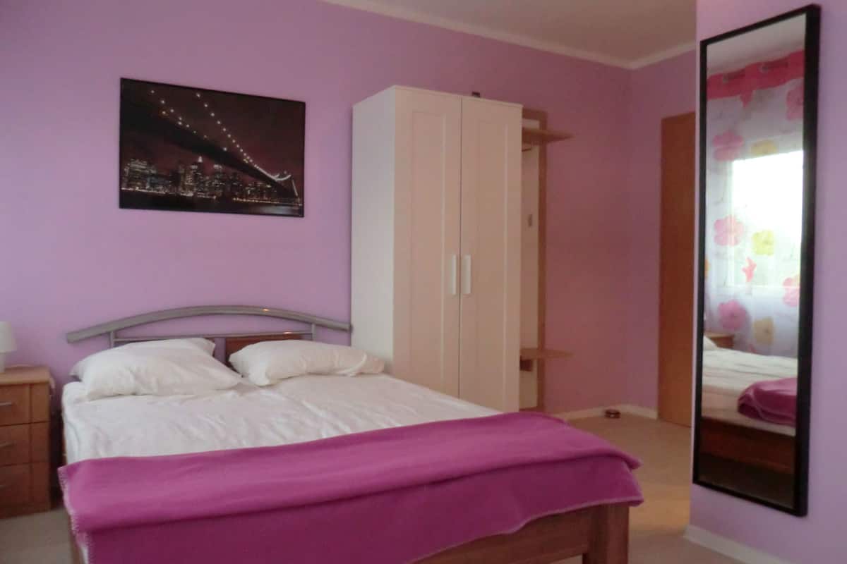 A bedroom with a bed, a wardrobe, a night cityscape painting, and a mirror reflecting a bed.