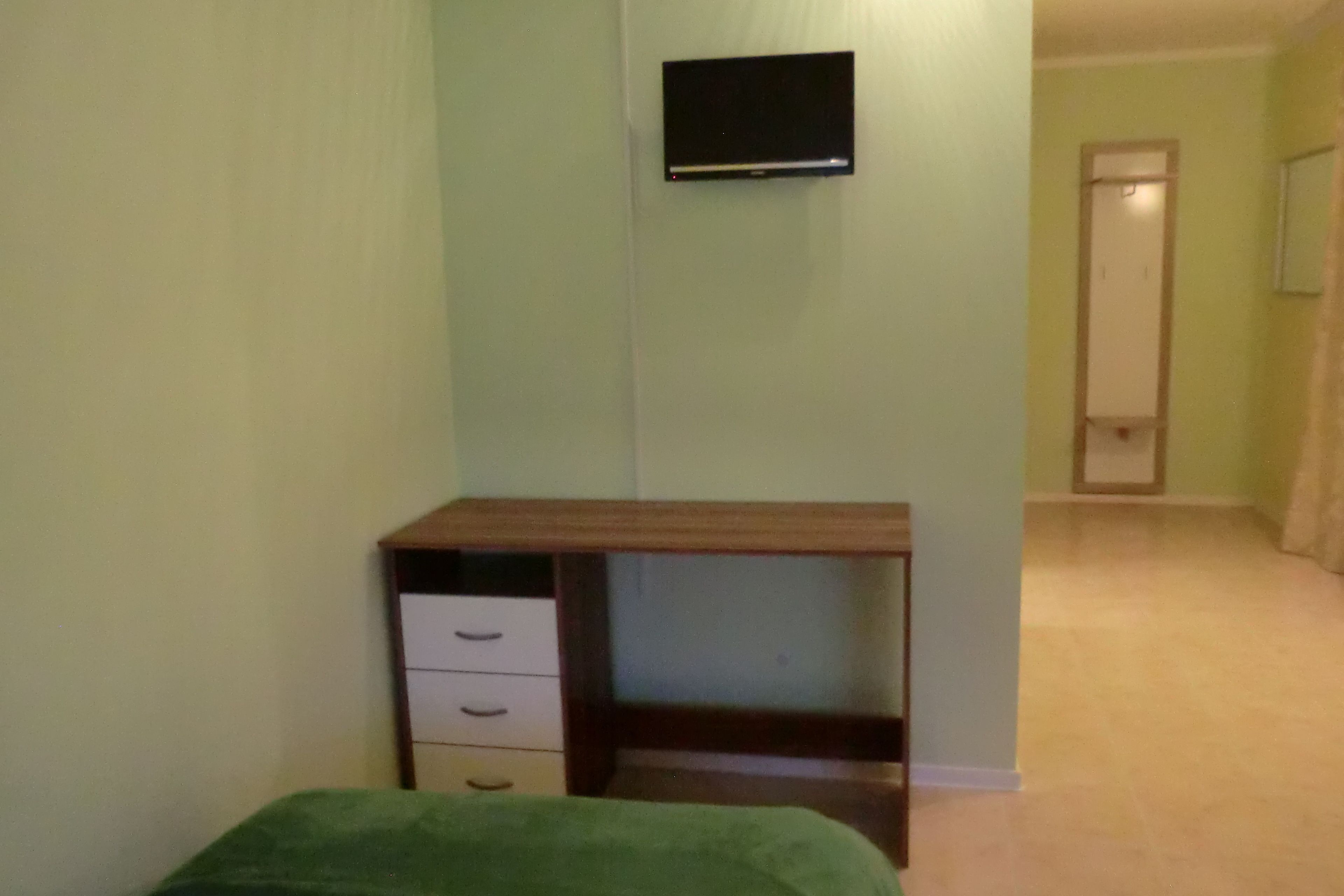 A room with a bed, a desk with drawers, a flat-screen TV mounted on the wall, and a door leading to another room.