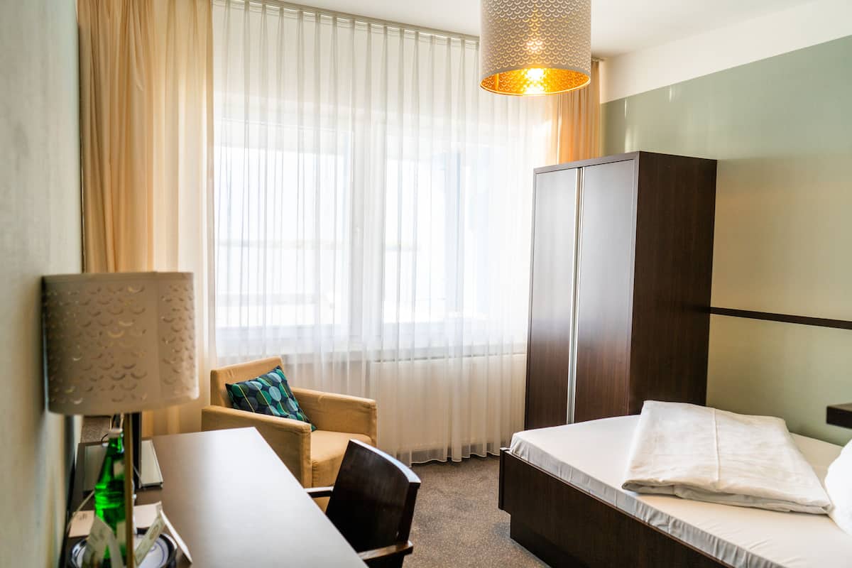 A hotel room with a bed, a desk, a chair, a wardrobe, and a window with sheer curtains.