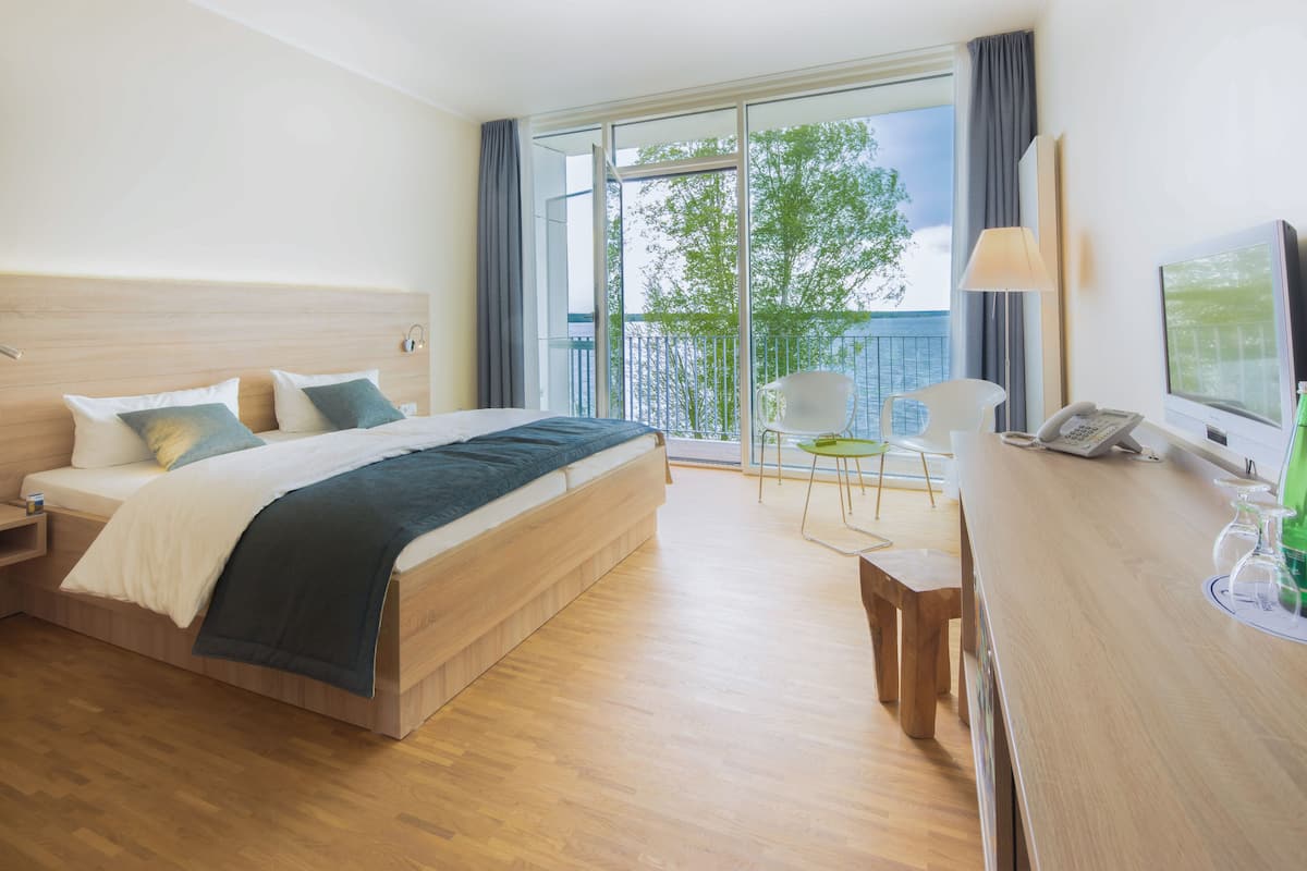 A hotel room with a large bed, a desk, a chair, a TV, and a balcony with a view.