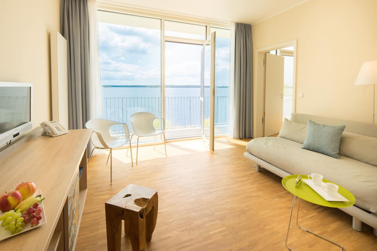 A hotel room with a wooden desk, a sofa, a balcony with a view of the sea, and a small table with cups and saucers.
