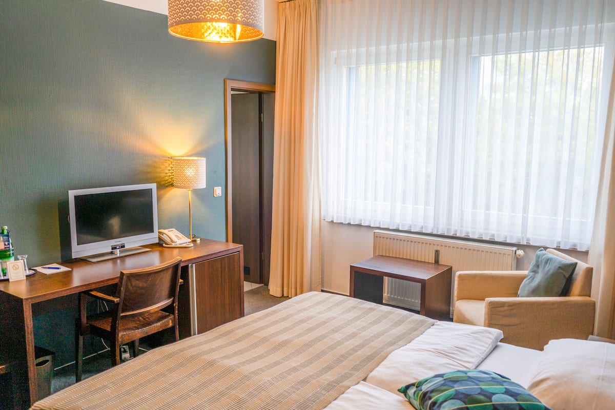 A hotel room with a bed, a desk with a television, a chair, a lamp, and a window with curtains.