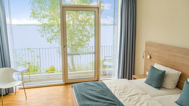 A bedroom with a large window, a bed with pillows, a chair, and a wooden headboard. - Strandhotel Senftenberger See (Senftenberg)