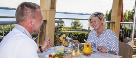 Ein Paar speist an einem Tisch im Freien mit Blick auf das Wasser, ein Korbstuhl und ein Tisch mit Essen und Getränken.