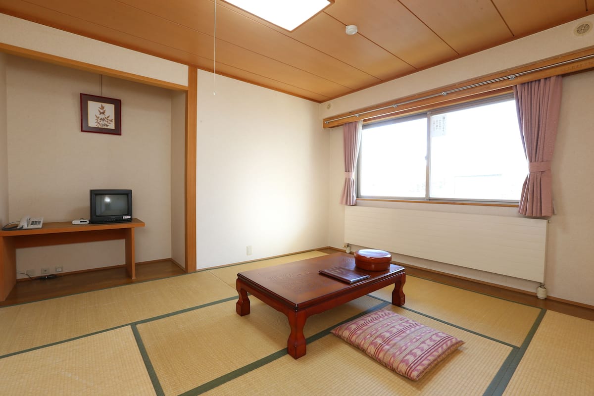 A traditional Japanese room with tatami flooring, a low wooden table, a television on a stand, and a window with curtains.