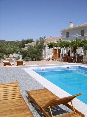 A swimming pool with wooden loungers, a covered seating area, and a white building with a tiled roof. - Lasnavillasmm (Montefrio)