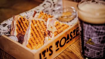 A wooden box labeled 'IT'S TOASTE' containing grilled sandwiches, a small bowl of sauce, and a glass of beer on a wooden table.