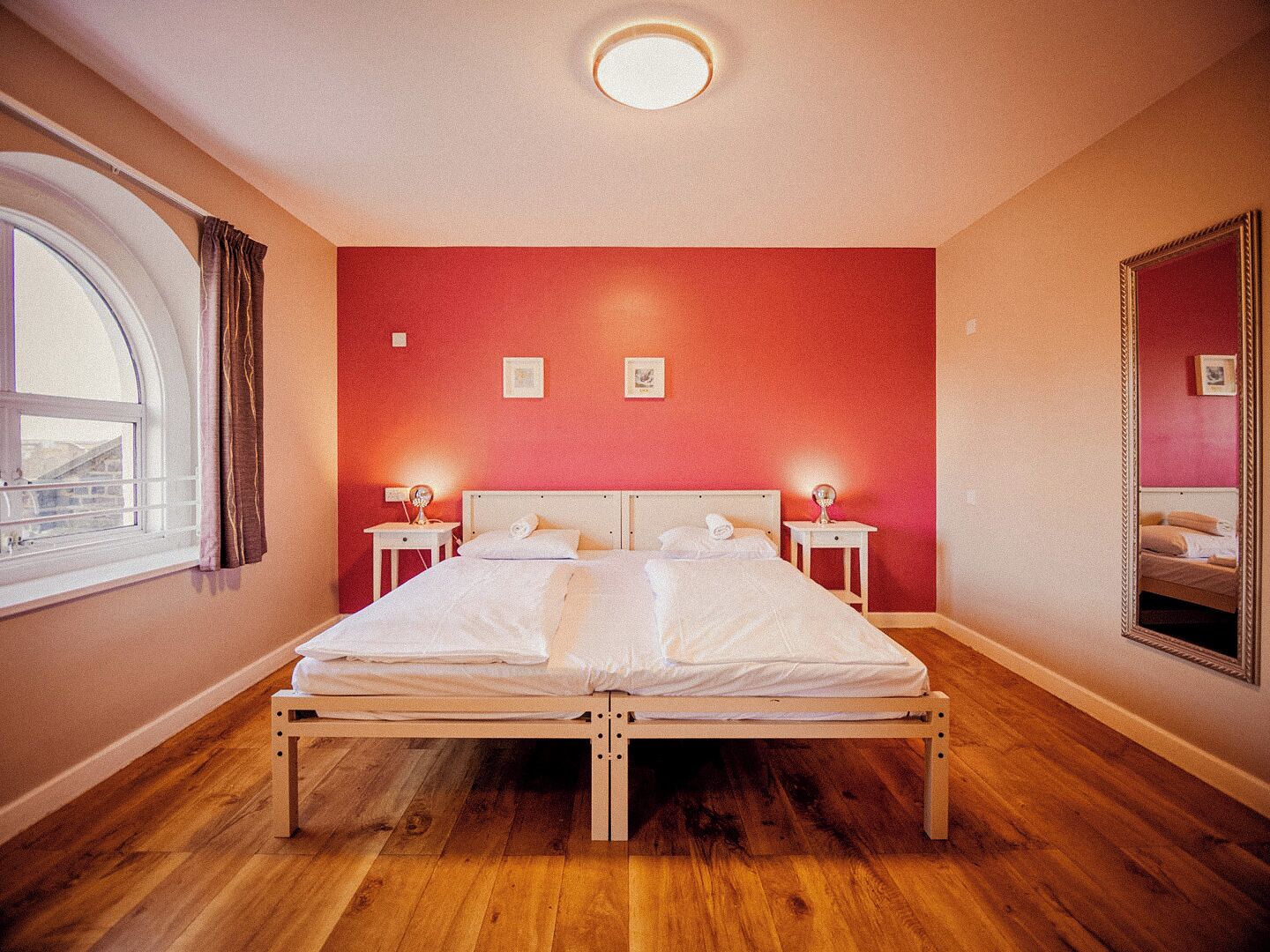 Une chambre à coucher avec un lit en bois, deux tables de chevet avec des lampes, un grand miroir et une fenêtre avec des rideaux.