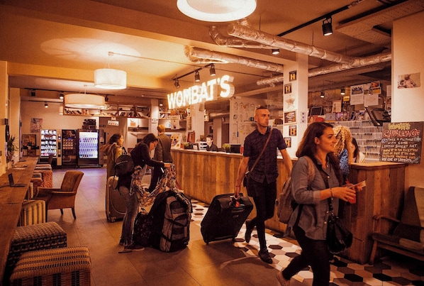 A bar area with a neon sign reading 'WOMBAT'S' and a counter with a menu. There are chairs and tables, and a refrigerator in the background. - Wombat's City Hostel London (London)