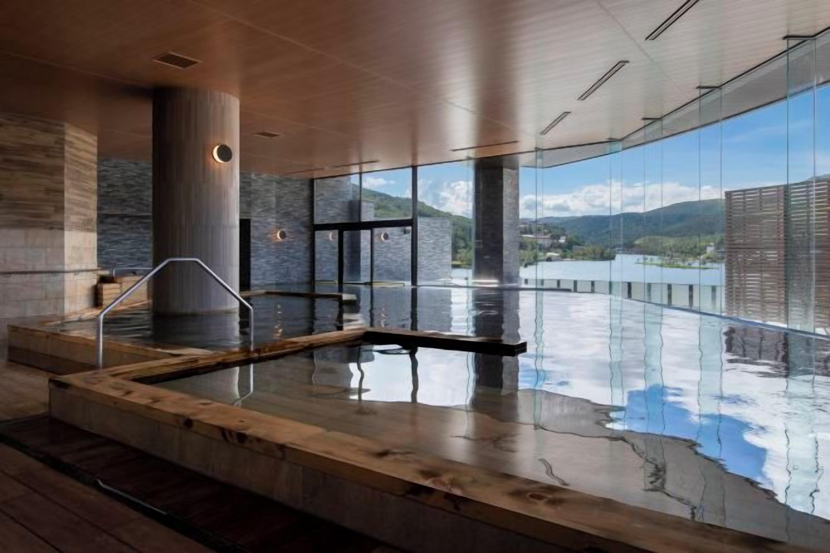 A modern indoor swimming pool with a wooden deck and glass walls offering a view of a lake and mountains.