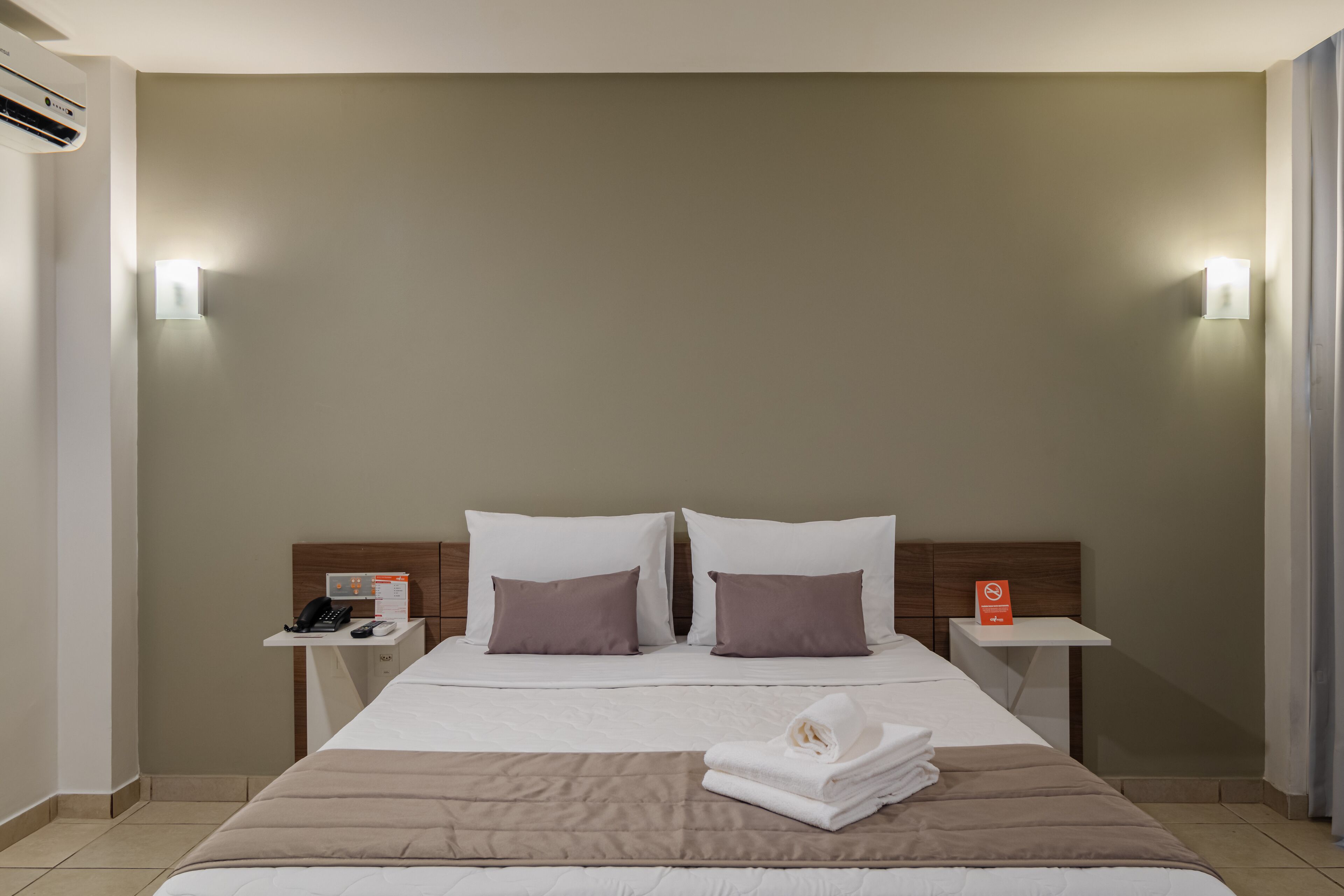 Ein Hotelzimmer mit einem großen Bett, zwei Nachttischen, einer Wandklimaanlage und einem Beistelltisch mit Telefon und Schild.