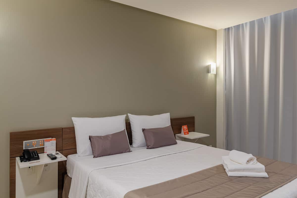 Een hotelkamer met een bed, een nachtkastje met telefoon en bedieningspaneel, een raam met gordijnen en een wandlamp.