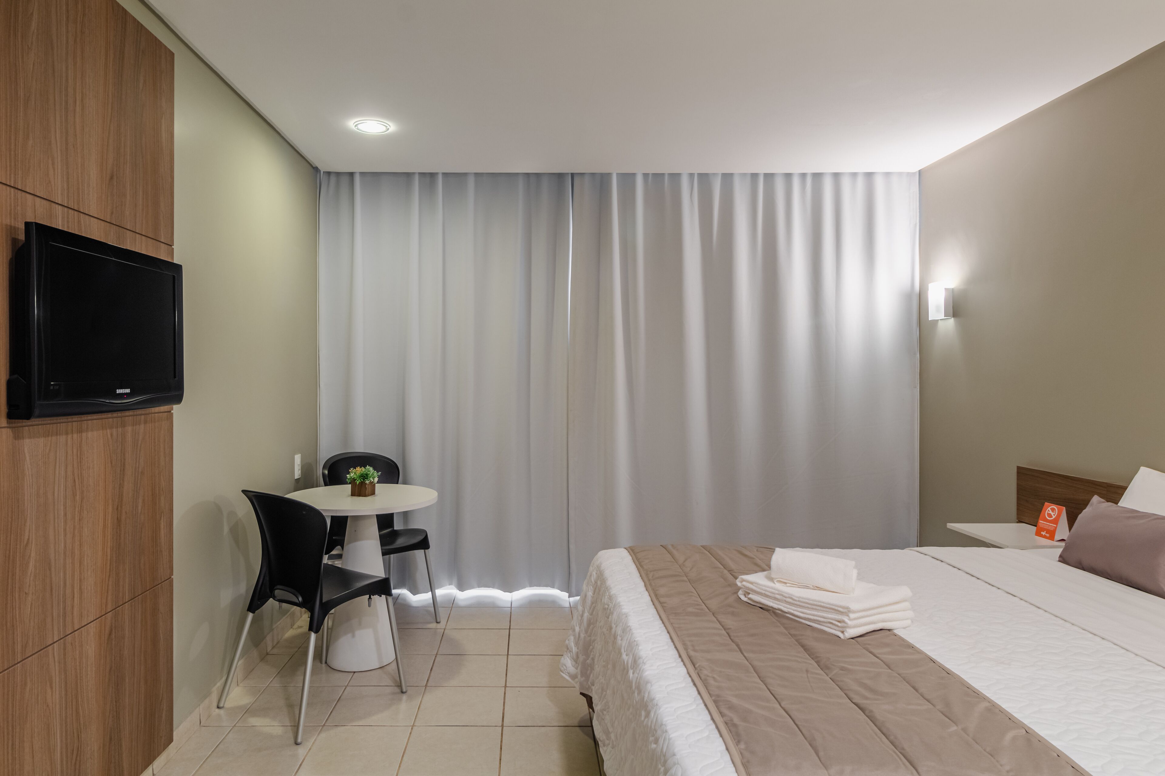 Una habitación de hotel con una cama, un escritorio con silla, un televisor fijado a la pared y una ventana con cortinas.