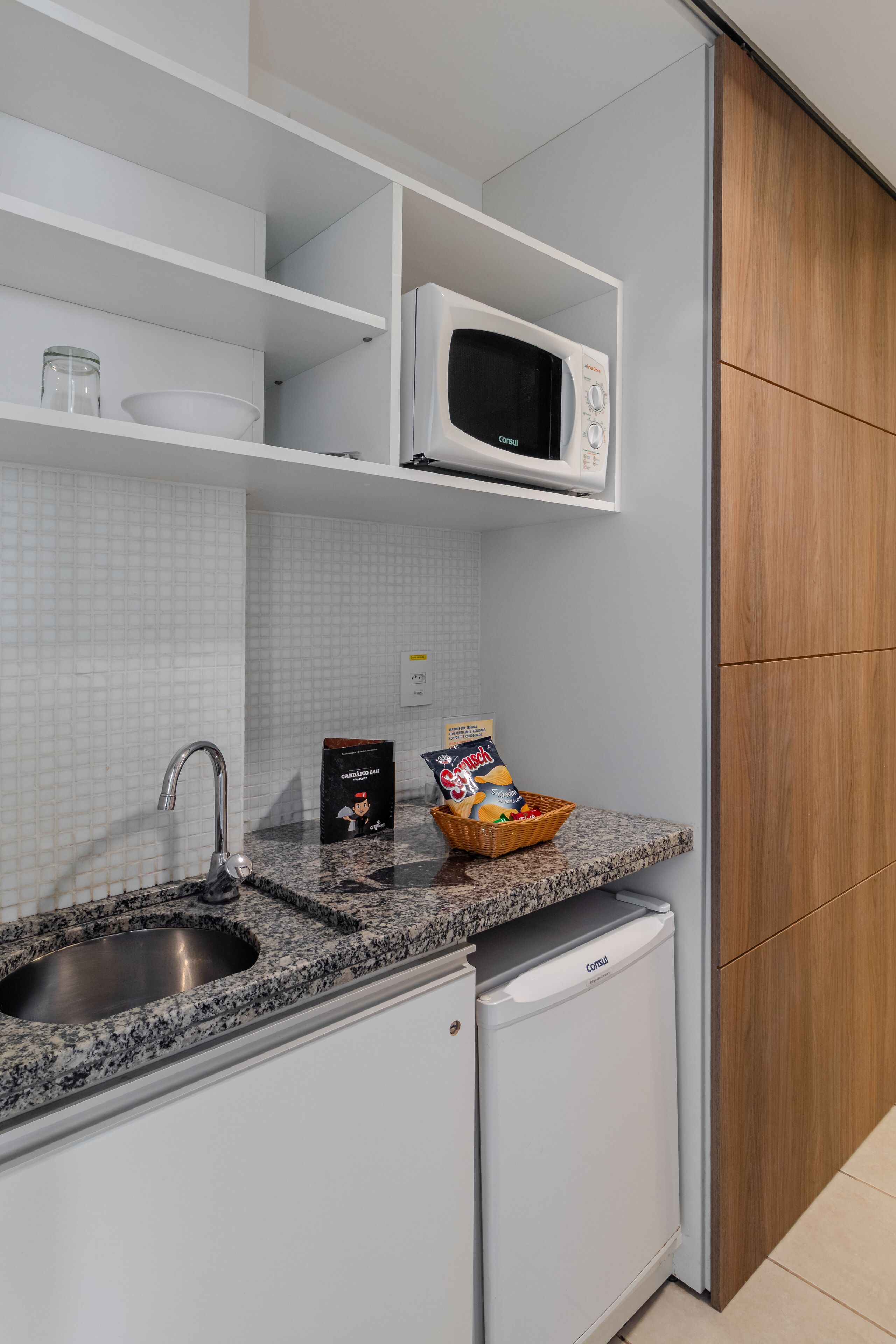 Una cocina con encimera de granito, pileta de acero inoxidable, microondas blanco y un refrigerador pequeño.