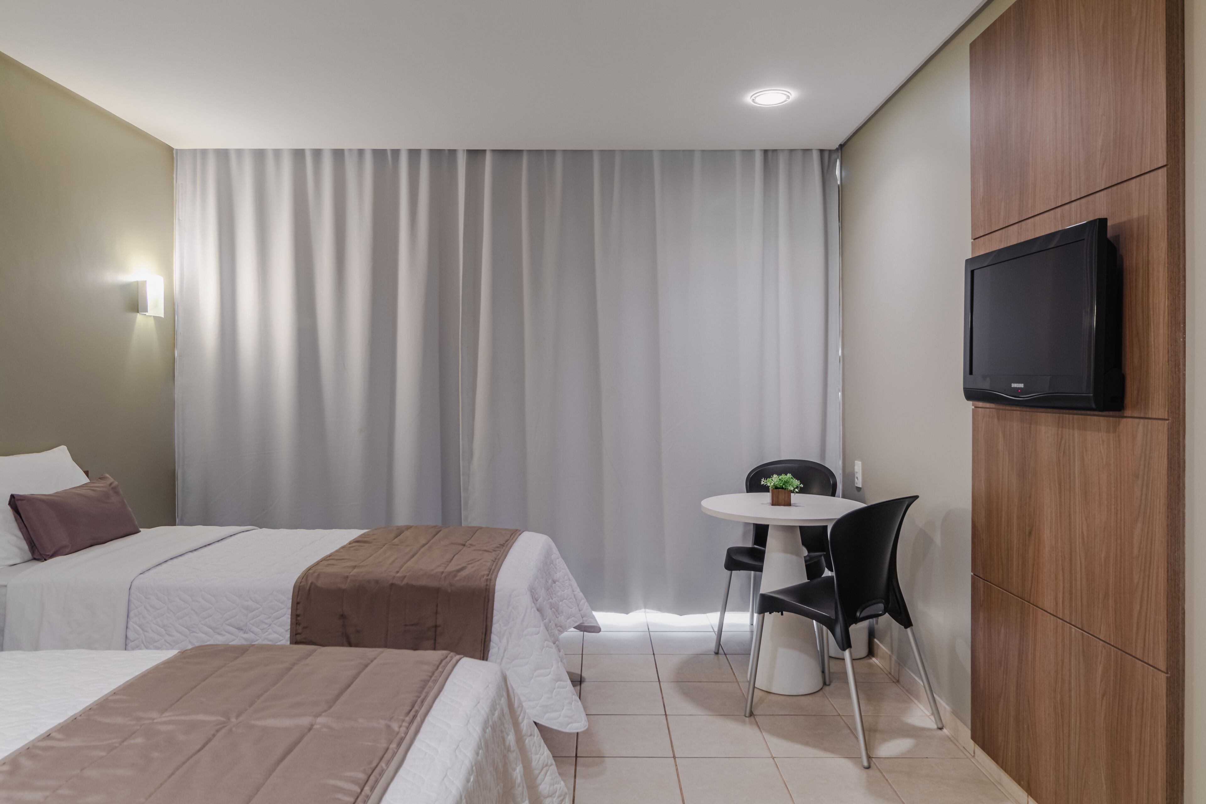 Een hotelkamer met twee bedden, een kleine tafel met stoelen, een aan de muur bevestigde televisie en een groot raam met gordijnen.