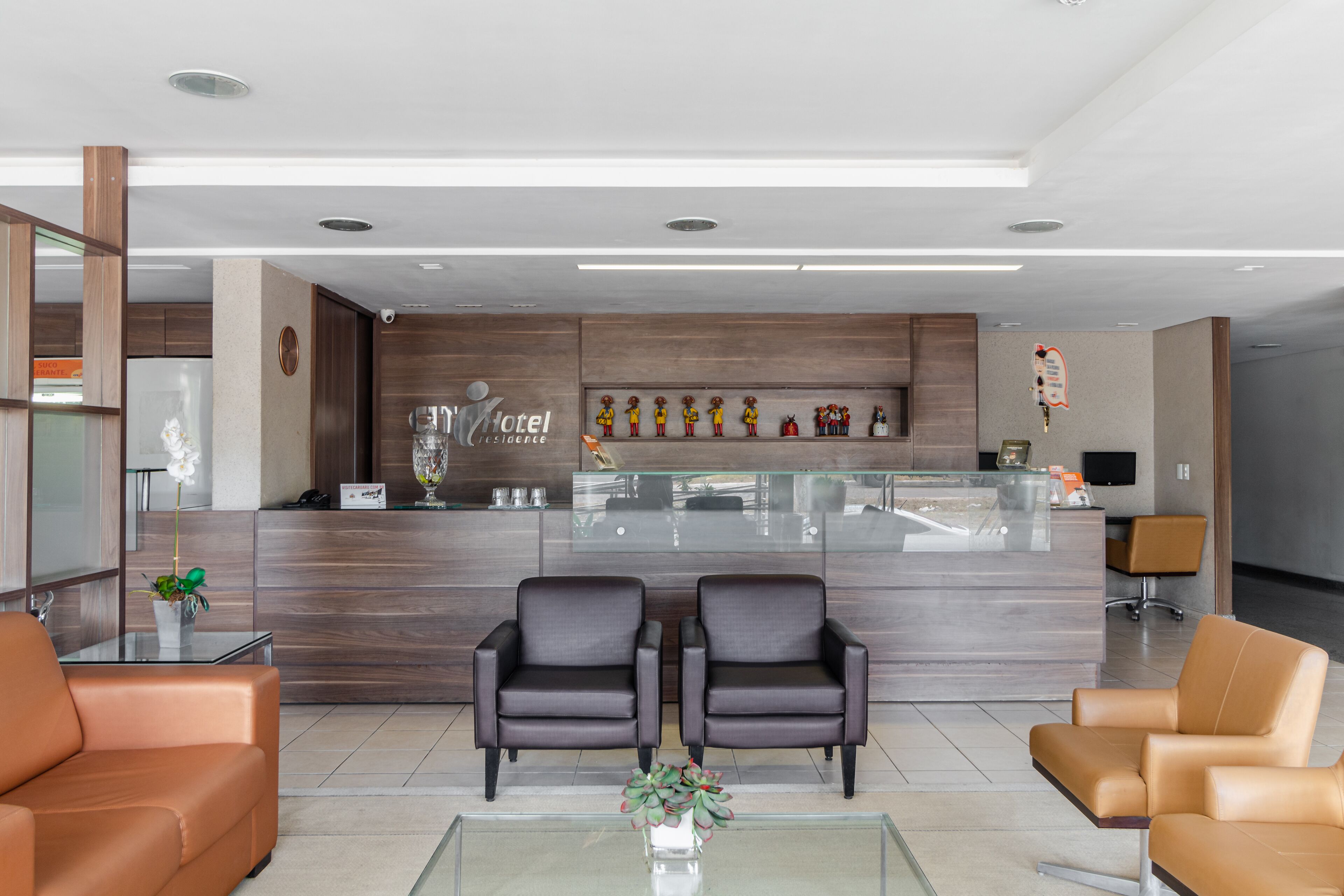 Un vestíbulo de hotel moderno con una recepción, dos sillones de cuero, una mesa de centro de vidrio y una planta decorativa.