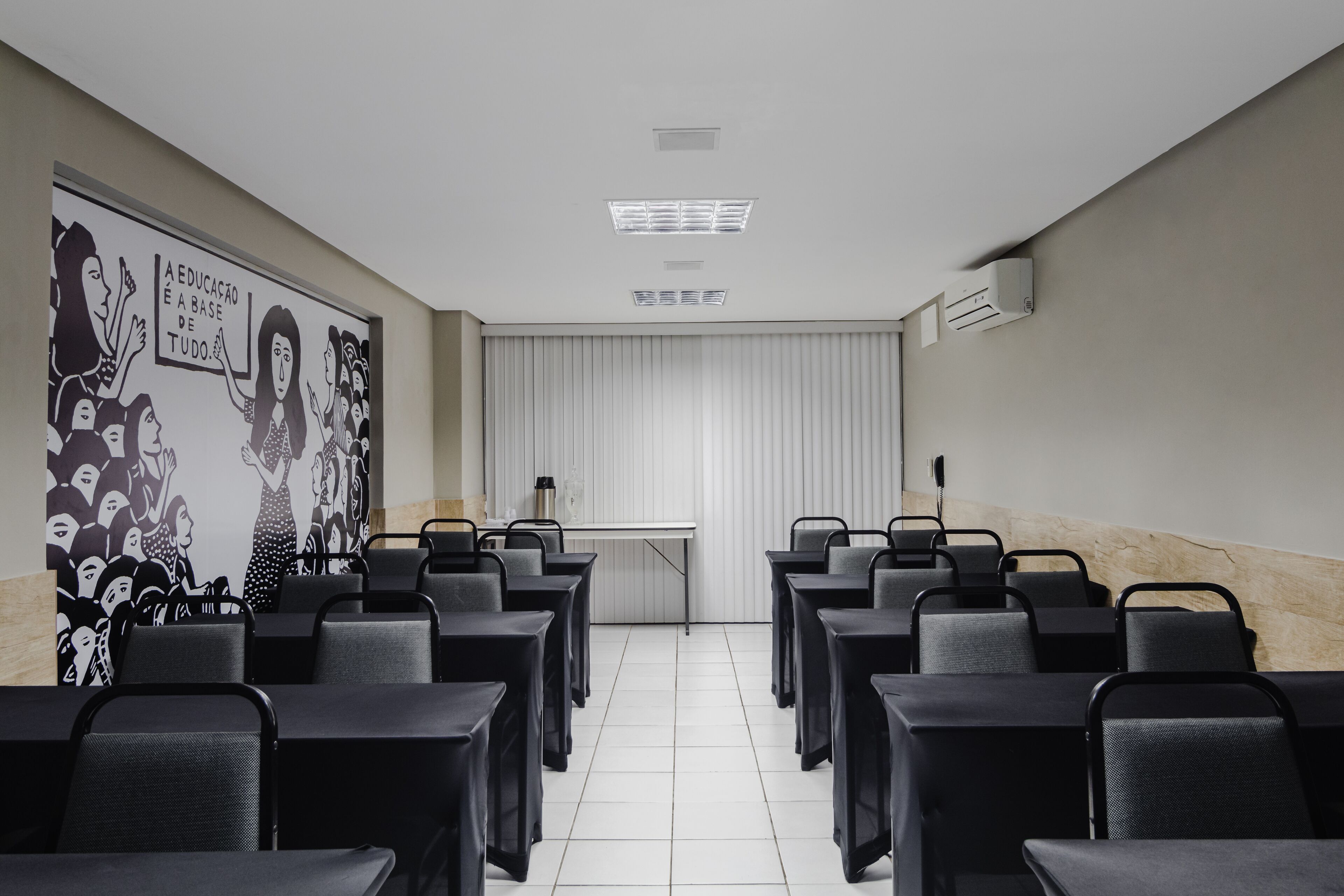 A conference room with rows of tables and chairs, a whiteboard with a black and white illustration, and a window with blinds.