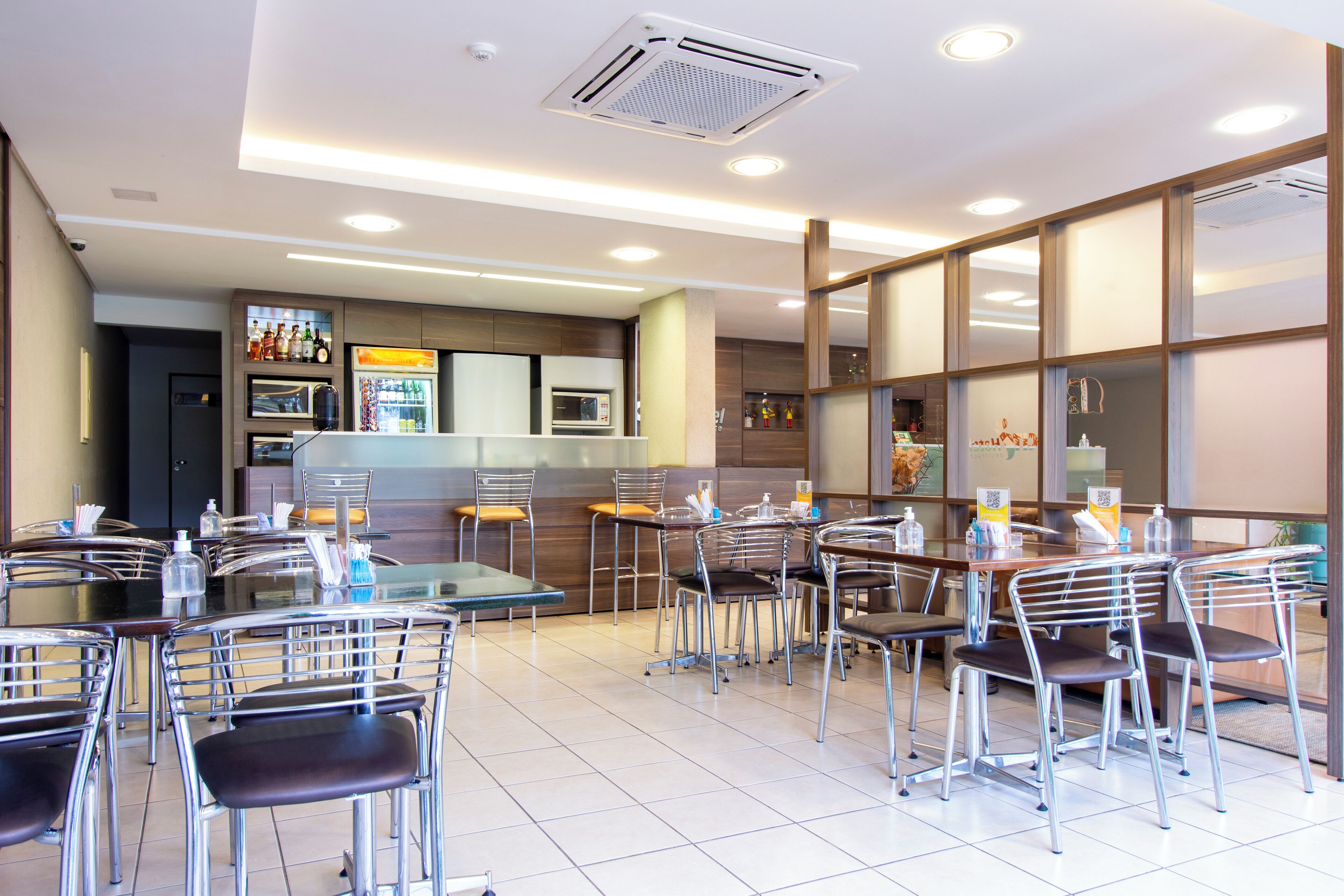 Un restaurante moderno con un interior limpio y minimalista, que presenta una mezcla de mobiliario metálico y de madera, una zona de comedor central y una cocina al fondo.