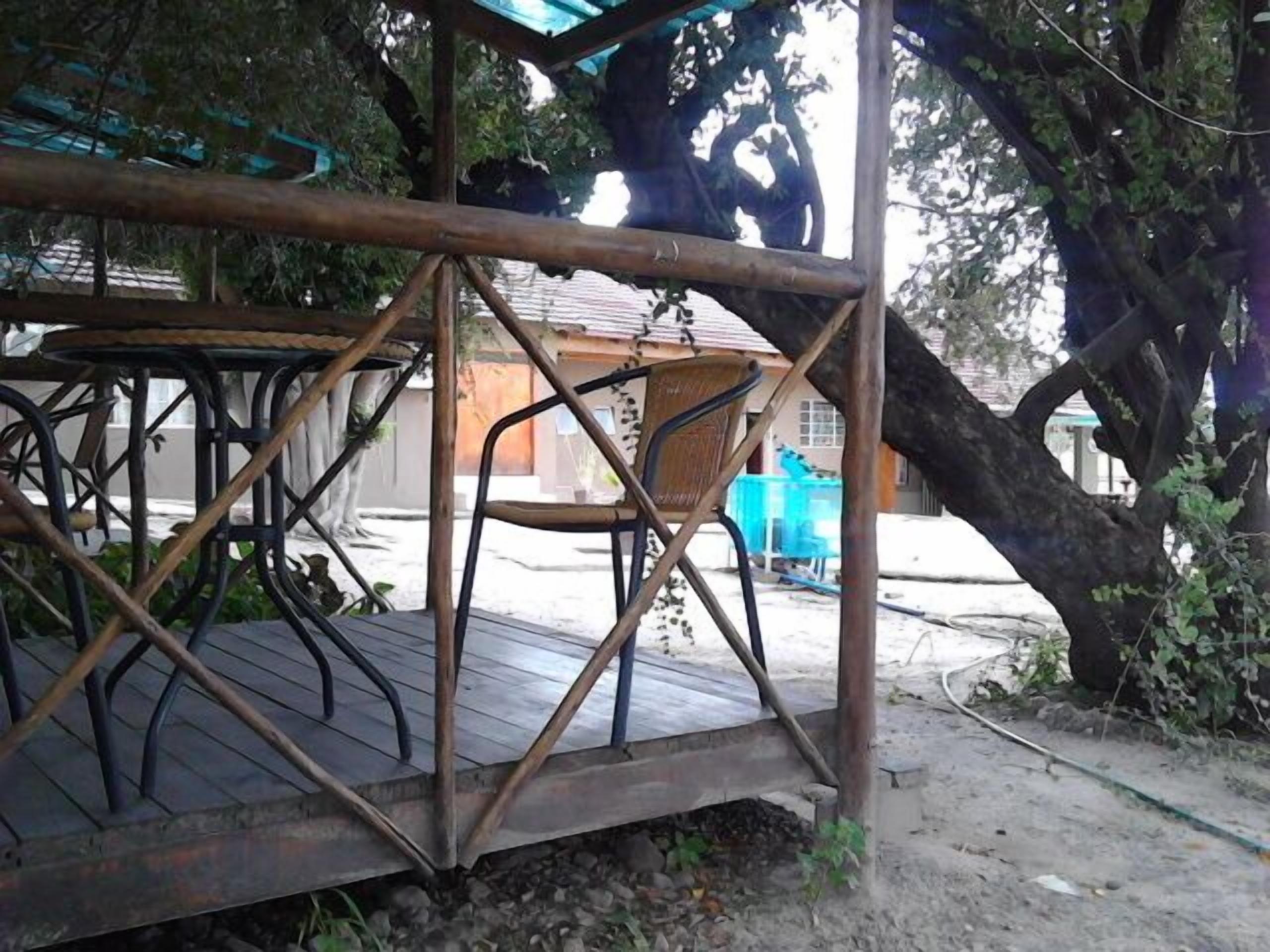 Un espace de détente extérieur de style rustique, doté d’une structure en métal et de planches de bois, entouré d’arbres et avec un bâtiment visible au loin.