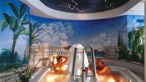Uma área de piscina interior luxuosa com duas banheiras de hidromassagem, um mural decorativo e uma garrafa de champanhe sobre uma mesa.