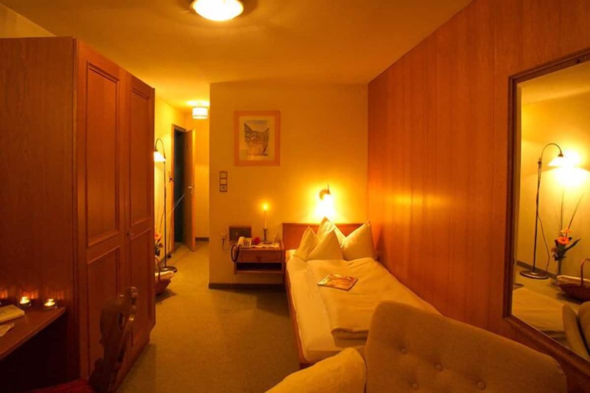 A hotel room with a bed, a chair, a desk with a lamp, a mirror, and a door leading to another room.