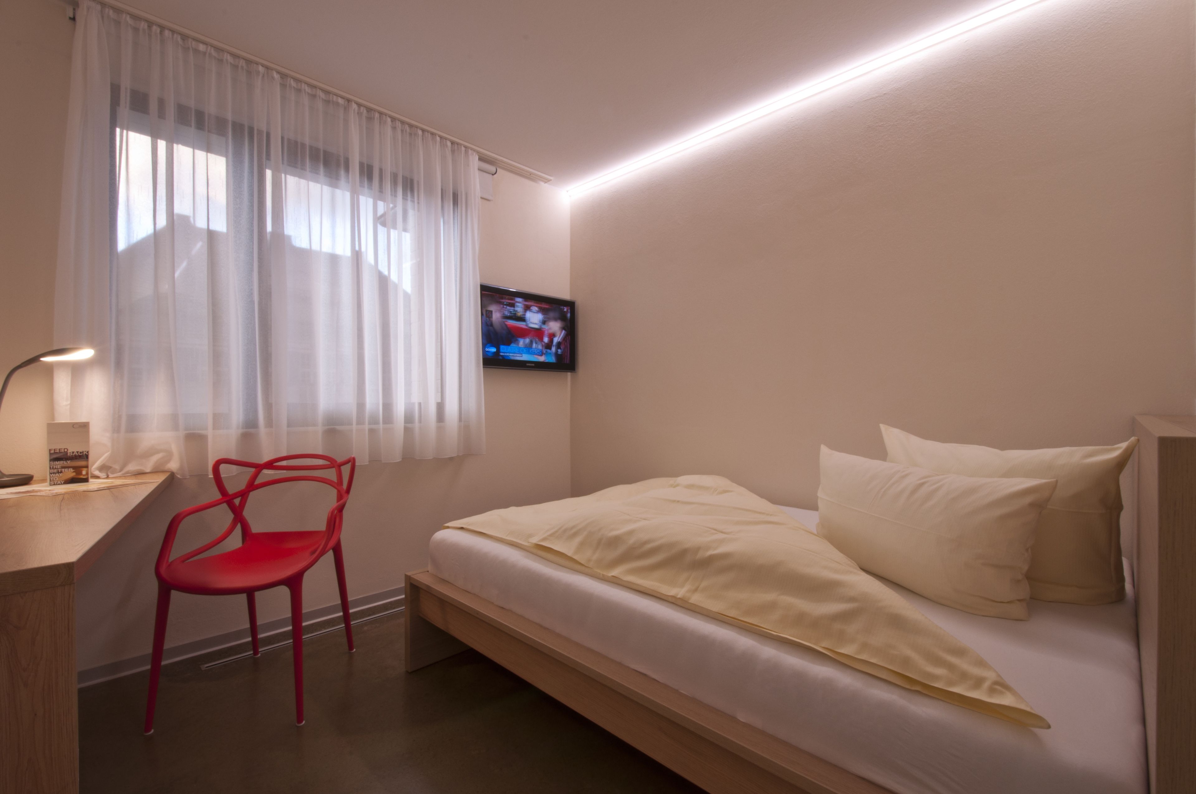 A bedroom with a bed, a desk with a chair, a TV, and a window with curtains.