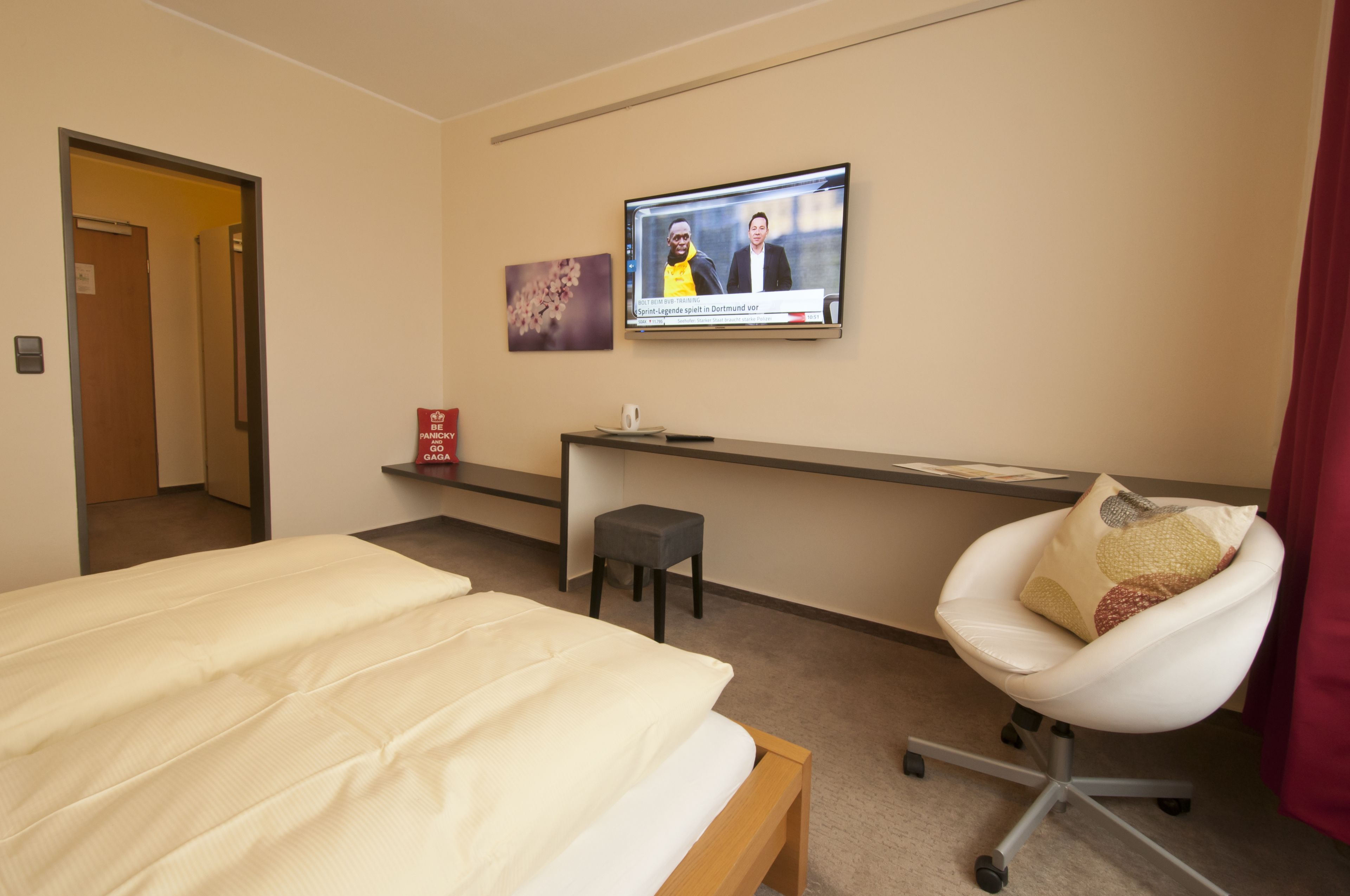 Una habitación de hotel con una cama, un escritorio con computadora, una silla y un televisor mostrando un programa de noticias.